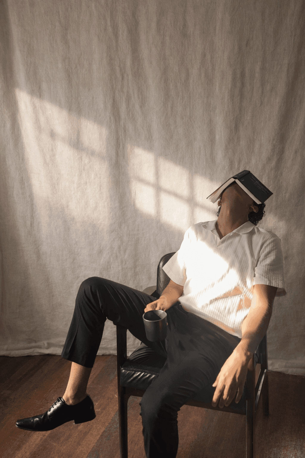 Matthew Arendse seated in a chair against a linen backdrop, an open book resting over his face, holding a cup of coffee in raking window light.