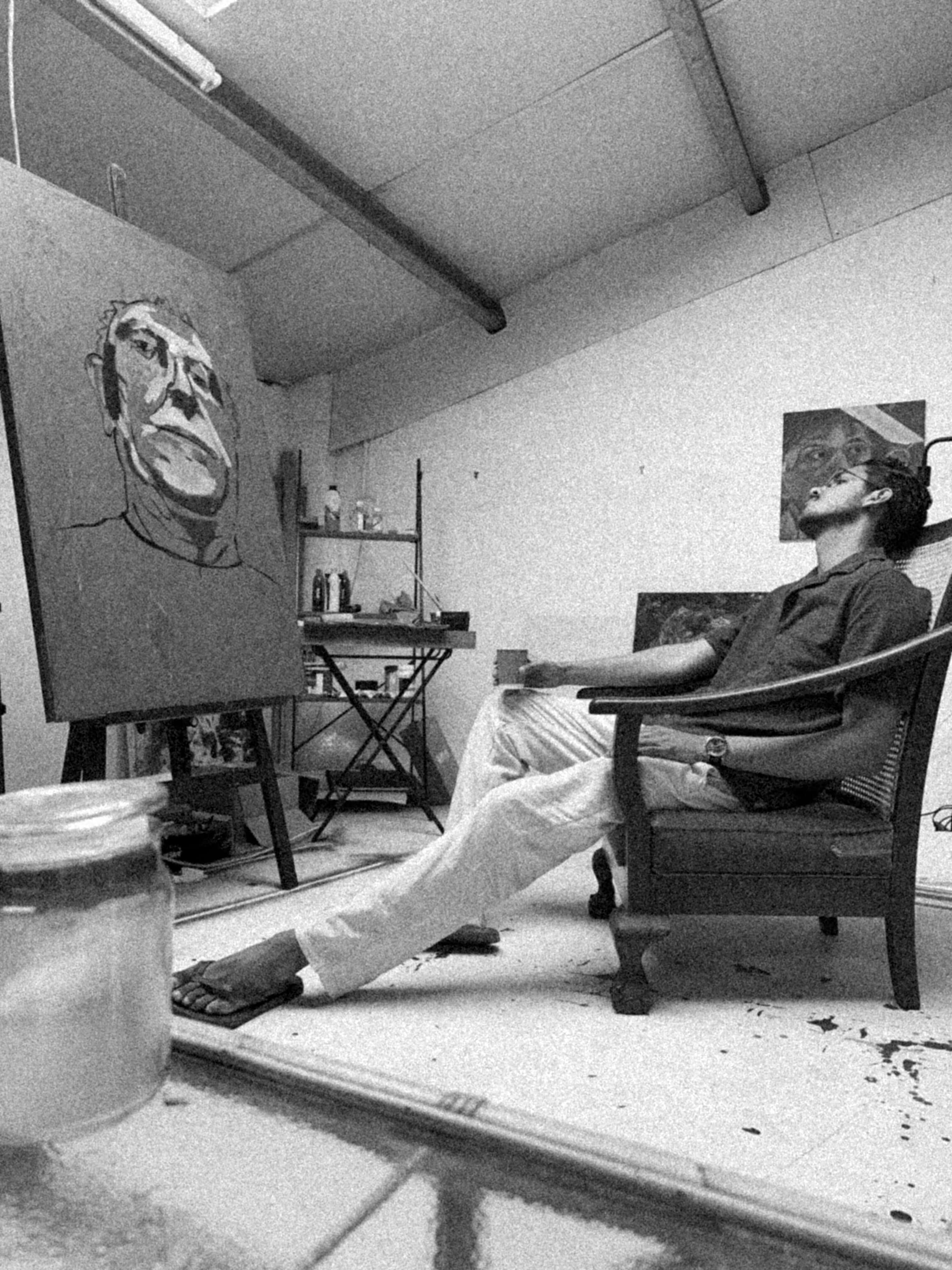 Matthew Arendse reclining in a chair in his studio, considering a large portrait in progress on the easel opposite him.